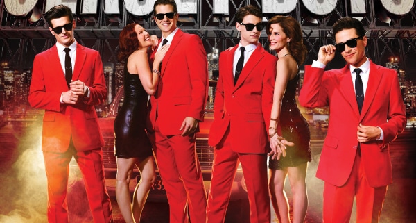 Jersey Boys. Four men in red suits and sunglasses accompanied by two doting women in black dresses. Fog effects and NYC skyline background