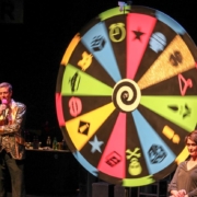Two actors appear beside a large, brightly-colored spinning wheel to emulate a Wheel of Fortune-type game