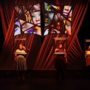 Two actors stand on stage. Each of their heads and shoulders stands just behind empty, suspended picture frames. The background is a multi-media projection of facial parts