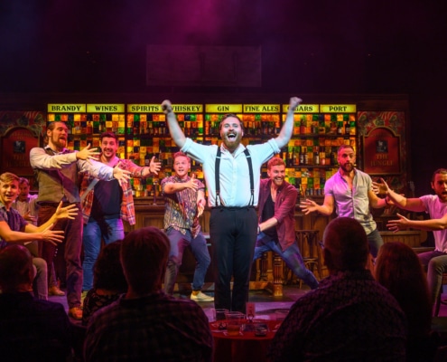 A bearded man wearing a white shirt, black trousers, and black suspenders raises his arms and sings towards the audience. He is at center stage, surrounded by other cast members who gesture towards him.