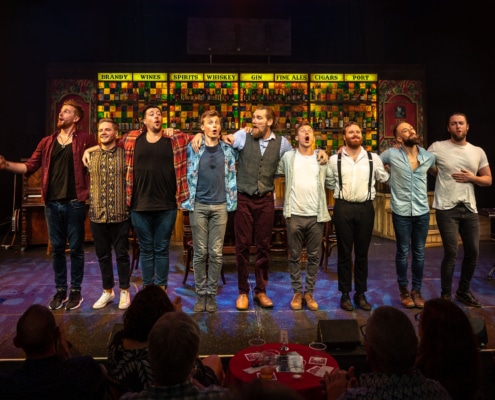 The nine primary cast members of The Choir of Man stand in a line, arm-in-arm, while singing towards the audience.