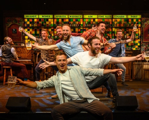 The nine primary cast members of The Choir of Man stand in a conga line, facing the audience, with their upper bodies gesturing in alternating directions. They hold musical instruments or glasses of beer in their hands.