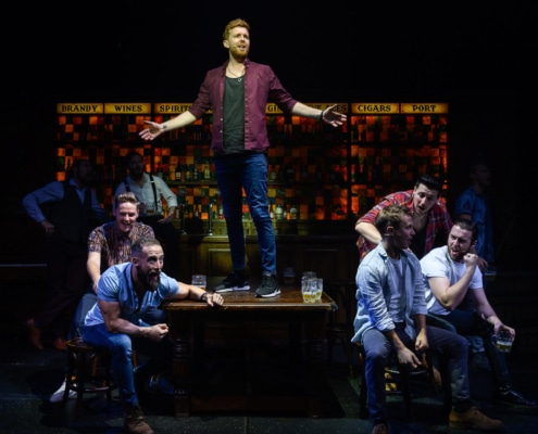 A cast member sings while standing on top of a table, surrounded by eight other cast members holding instruments and glasses of beer.