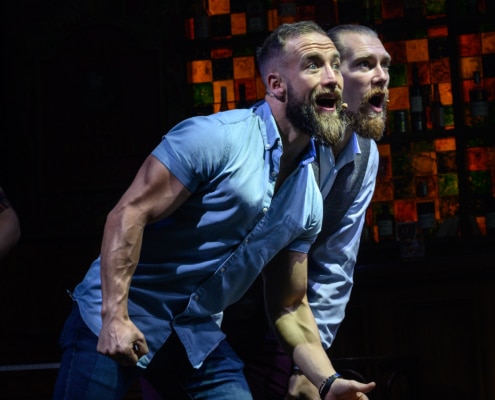 Two bearded cast members wearing blue shirts pose and sing on stage.