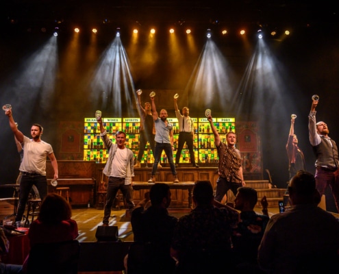 The nine primary cast members of The Choir of Man stand in formation while raising empty beer glasses upward with their right hands.