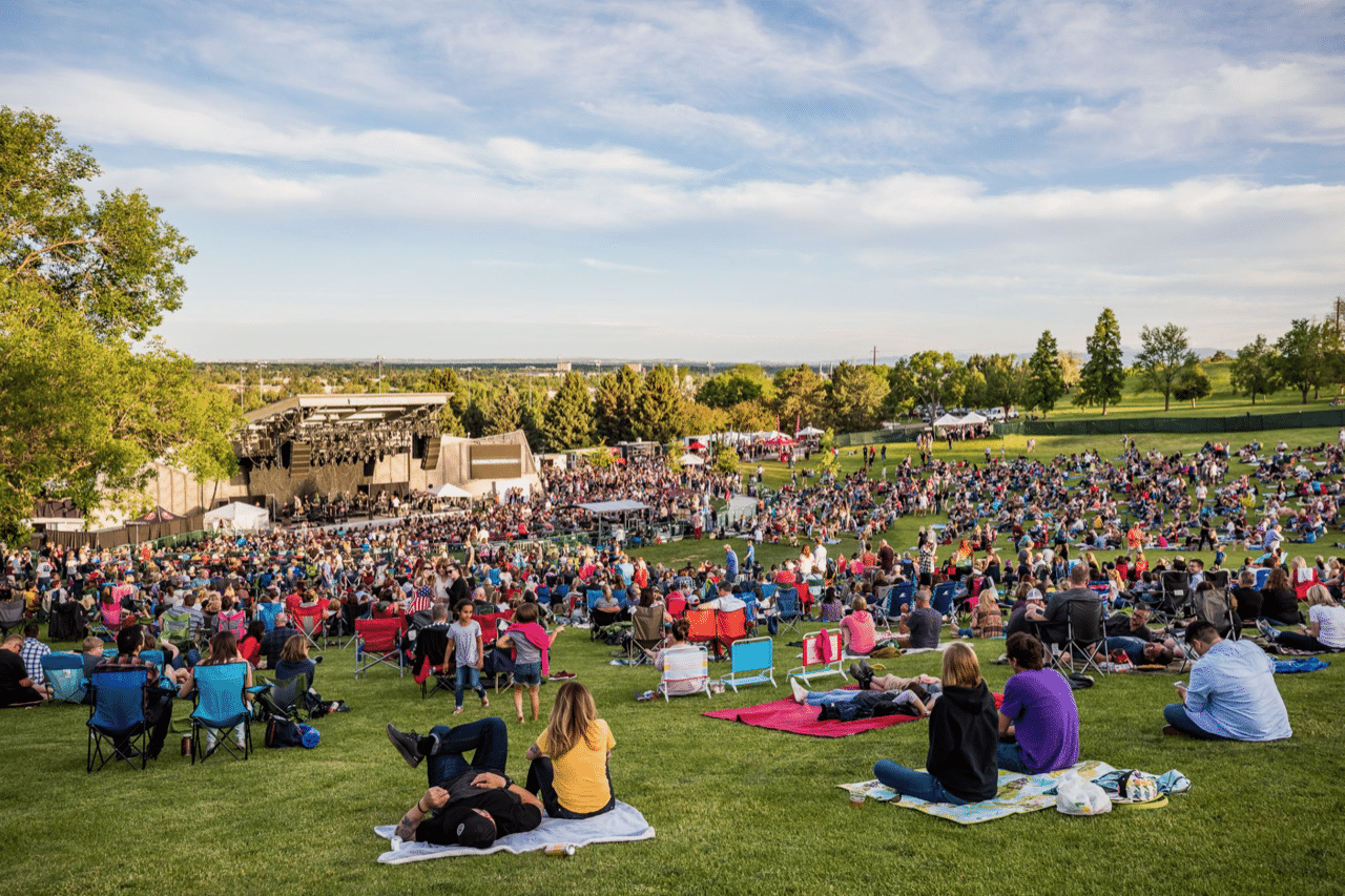 A crowd of people sitting on blankets in the grass, facing an amphitheatre in the distance, while the sky is blue with gentle clouds.