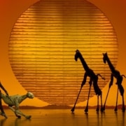 Puppets of a cheetah and two giraffes are silhouetted against a setting sun backdrop.