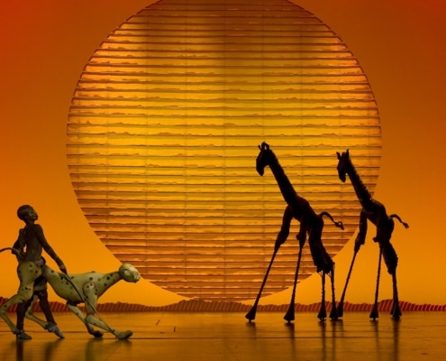 Puppets of a cheetah and two giraffes are silhouetted against a setting sun backdrop.