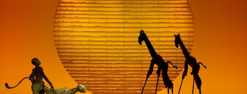 Puppets of a cheetah and two giraffes are silhouetted against a setting sun backdrop.