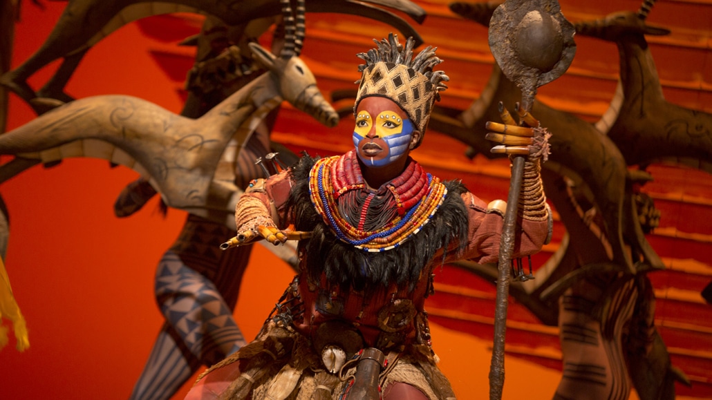 A person dressed in traditional African attire with a headdress and colorful beaded necklaces, holding a staff, performing in front of an orange background with animal figures.