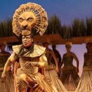 A theatrical performance featuring a performer in an elaborate costume with a lion headpiece, surrounded by other performers in traditional attire. The background includes tall grass and a blue sky, creating an African savanna setting.