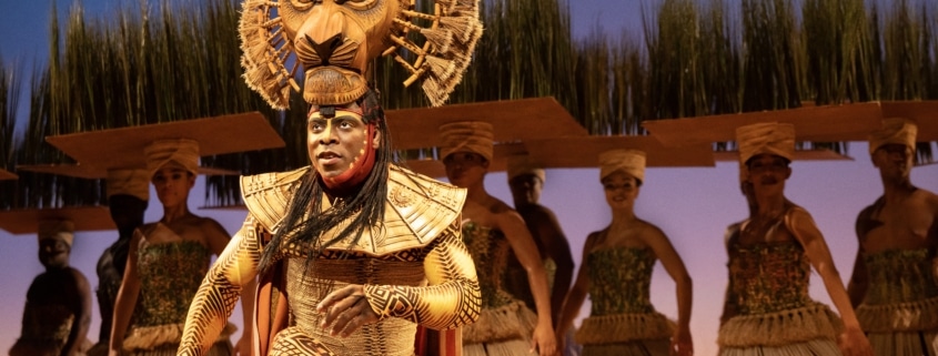 A theatrical performance featuring a performer in an elaborate costume with a lion headpiece, surrounded by other performers in traditional attire. The background includes tall grass and a blue sky, creating an African savanna setting.