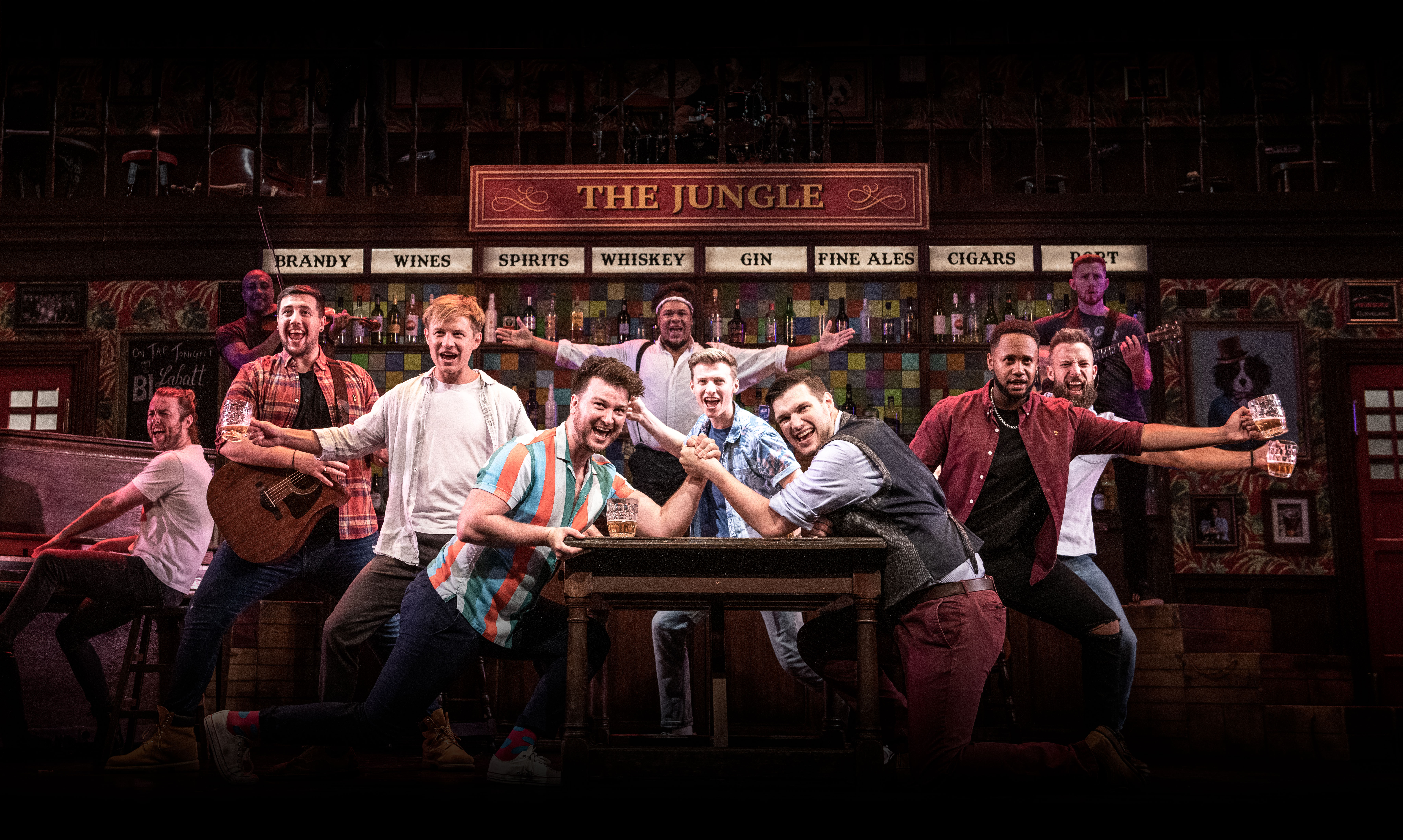 Two men arm wrestle on a pub table as they smile and sing. Nine other cast members pose with their arms stretched out, some with glasses of beer or instruments in their hand.