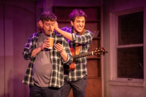 Actor Seth Dhonau places his hands over actor Brett Ambler’s ears and grins while Ambler sips a drink from a straw and looks at a smartphone