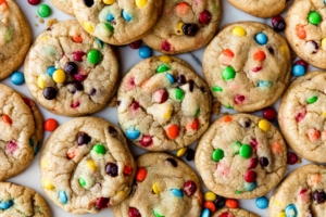 Rainbow M&M cookies arranged in slightly overlapping rows with loose M&M candies on a white surface