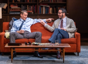 two men sit on a red couch pointing their fingers at each other