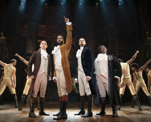 Cast of Hamilton Broadway National Tour poses in front of dancers in background onstage