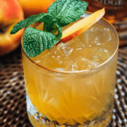 a rocks glass filled with a yellow beverage and topped with a peach slice and mint leaves is sitting in a brown wicker surface with whole peaches in the background