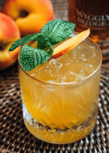 a rocks glass filled with a yellow beverage and topped with a peach slice and mint leaves is sitting in a brown wicker surface with whole peaches in the background