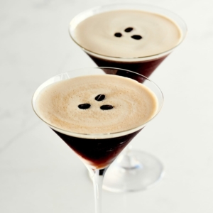 two martini glasses on a white countertop filled with a brown beverage and topped with three coffee beans each