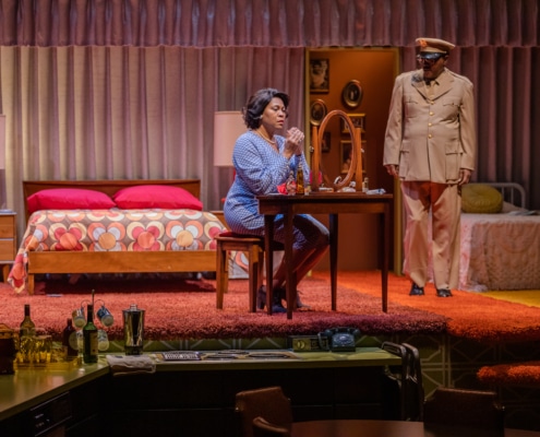 Chavez Ravine sits at a vanity as Levy Lee Simon stands, in military dress, on stage during a performance of In The Upper Room