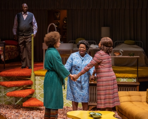 Levy Lee Simon stands at the top of a staircase and looks down at Sydney Cole Alexander, Yvette Monique Clark, and Monnae Michaell, who are standing in a circle, holding hands, and praying