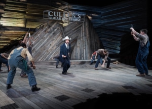 Matthew Bryan Feld appears on stage wearing a pinstripe suit and cowboy hat, surrounded by other actors who gesture towards him