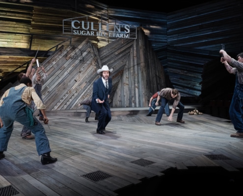 Matthew Bryan Feld appears on stage wearing a pinstripe suit and cowboy hat, surrounded by other actors who gesture towards him