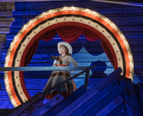 Leana Rae Concepcion, as Annie Oakley, slings a prop rifle over her shoulder while standing in the center of a circle of light bulbs