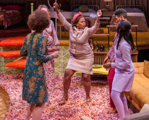 Monnae Michaell and the cast of In the Upper Room, dressed in their Sunday best, dance in a circle in a 70s-themed living room setting, complete with pink shag carpet