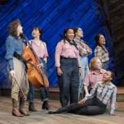 Seven female cast members, including Pekarek, pose and sing on a stage made of distressed wood in front of a blue backlight