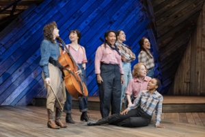 Seven female cast members, including Pekarek, pose and sing on a stage made of distressed wood in front of a blue backlight