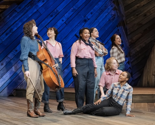 Seven female cast members, including Pekarek, pose and sing on a stage made of distressed wood in front of a blue backlight