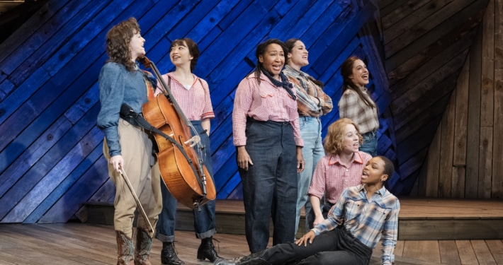 Seven female cast members, including Pekarek, pose and sing on a stage made of distressed wood in front of a blue backlight