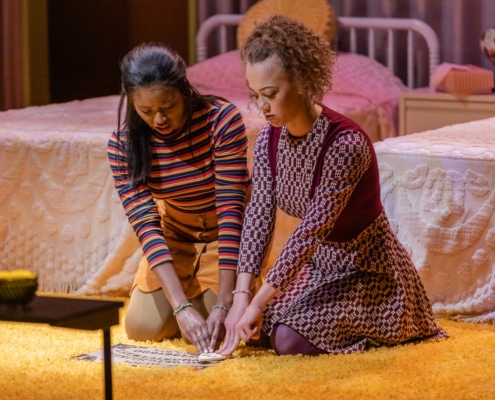 Courtney A. Vinson, as Josephine, and Kayla King, as Yvette, kneel side-by-side on a carpeted floor and guide the planchette of a Ouija Board together