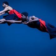 CPRD Ensemble dancers leap in front of a blue background wearing black and red costumes