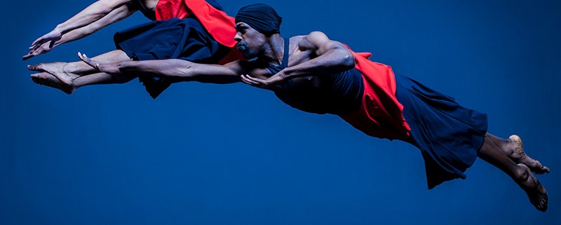 CPRD Ensemble dancers leap in front of a blue background wearing black and red costumes