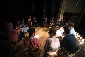 actors sit in a circle on folding chairs reading from scripts