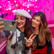 Two women pose for a selfie. One is wearing a white beret. Both are in front of a vivid pink background