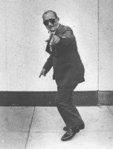 Black and white photo of a man in a suit and sunglasses pointing at the camera