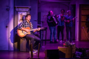 Narrator Josh of The Other Josh Cohen strums a guitar and sings into a microphone