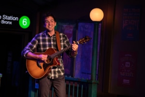 Narrator Josh of The Other Josh Cohen holds a guitar onstage