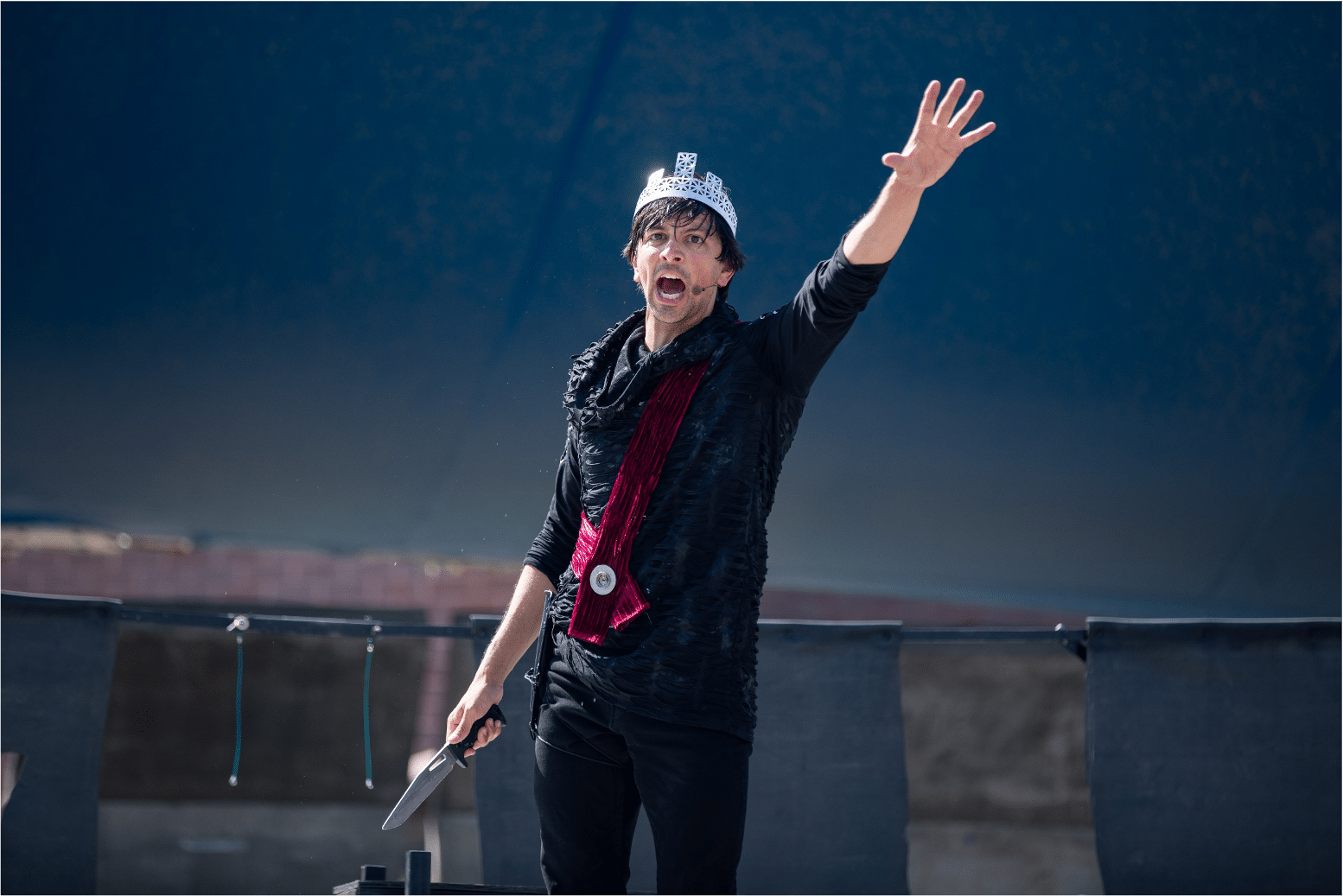 Man in all black, a red sash, and silver crown yells out towards the audience with a dagger in his right hand