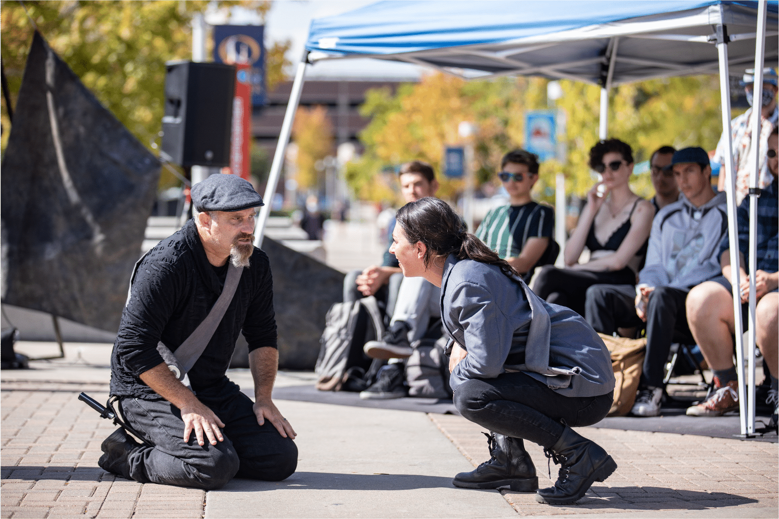 Actor in silver sash and newsboy cap on his knees conversing with actress in a silver blazer and sash crouching to his level