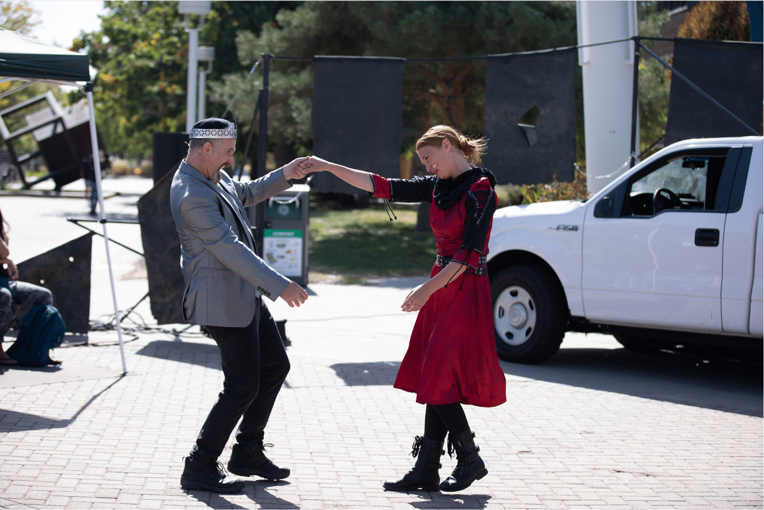 Man in silver blazer and crown dances happily with lady in a ruby red dress next to white pickup truck