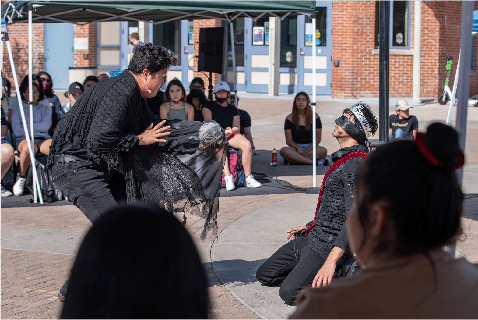 Actor holds a human skull under a sheer black sheet towards a blindfolded man on his knees in a red sash and silver crown