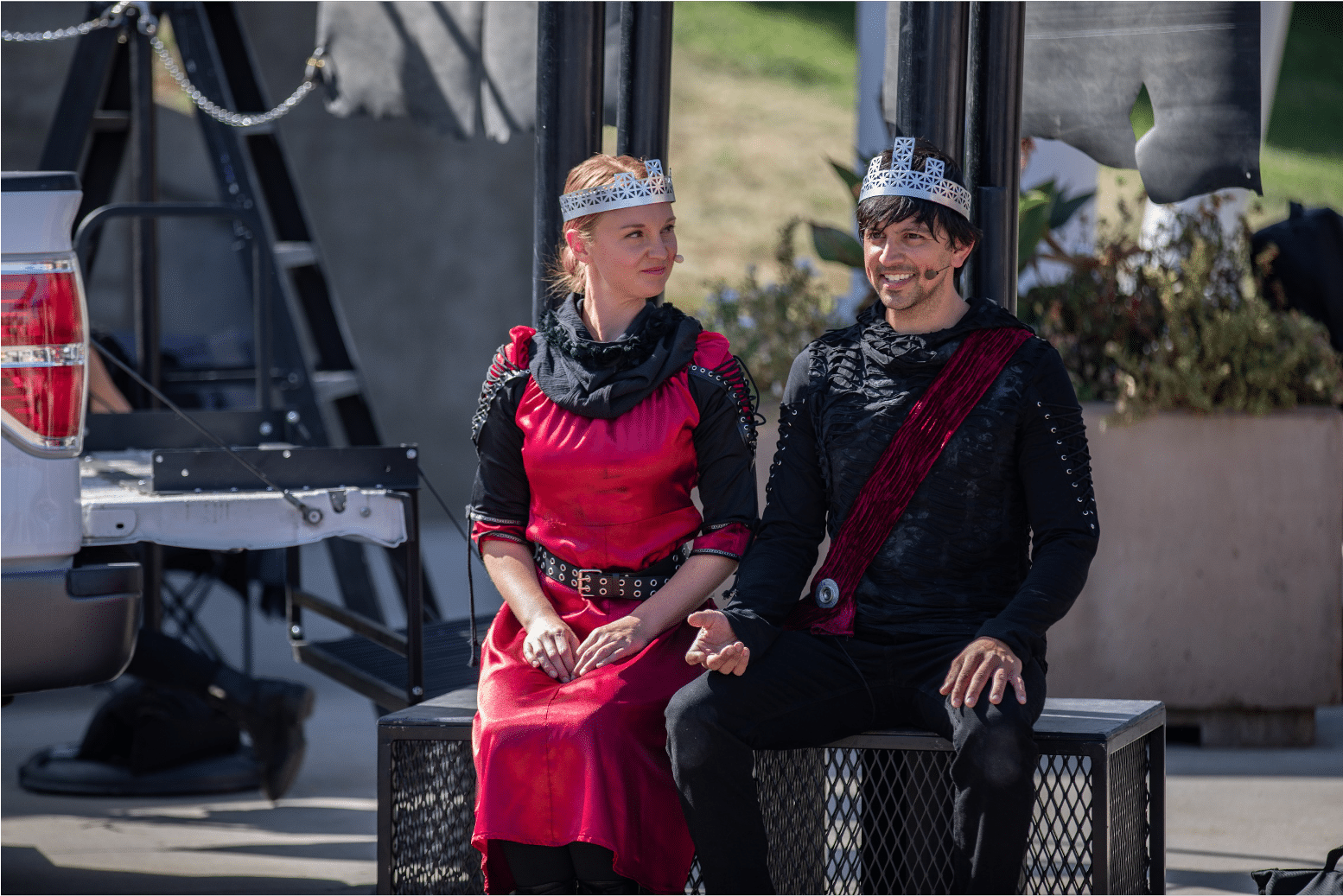 Lady in red sits next to man in red sash. Both wear silver crowns. She looks towards him as he looks outward, both with pride