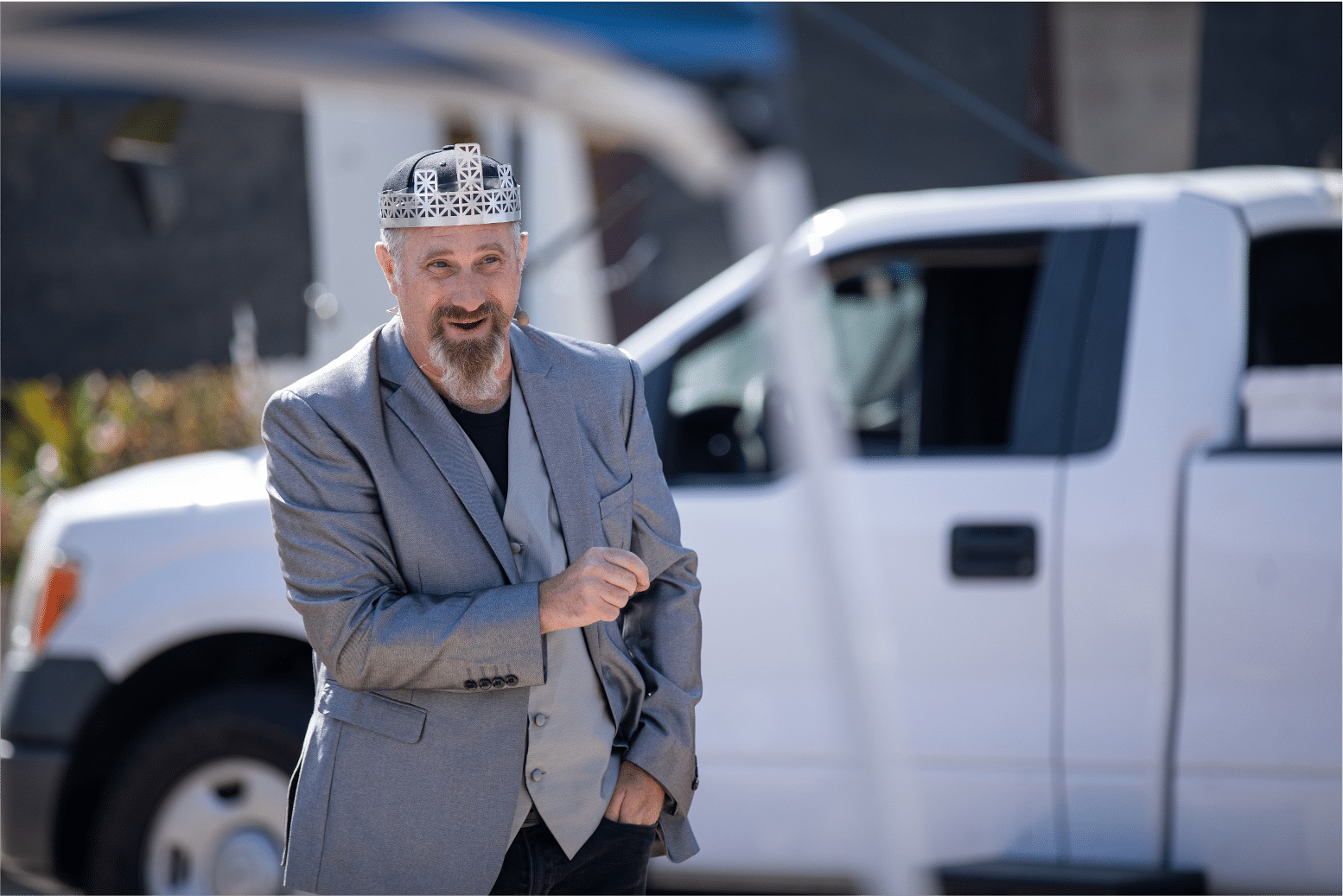 Man in silver blazer and crown speaks to the audience. White pickup truck blurred behind him.