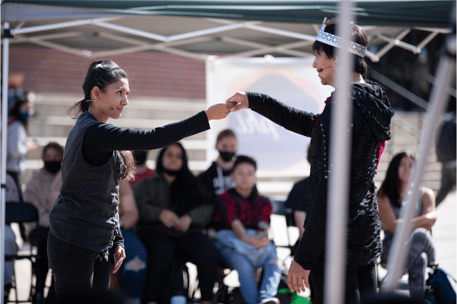 Actress in grey sweater holds up the hand of the actor in a red sash and silver crown as she looks at him with pride