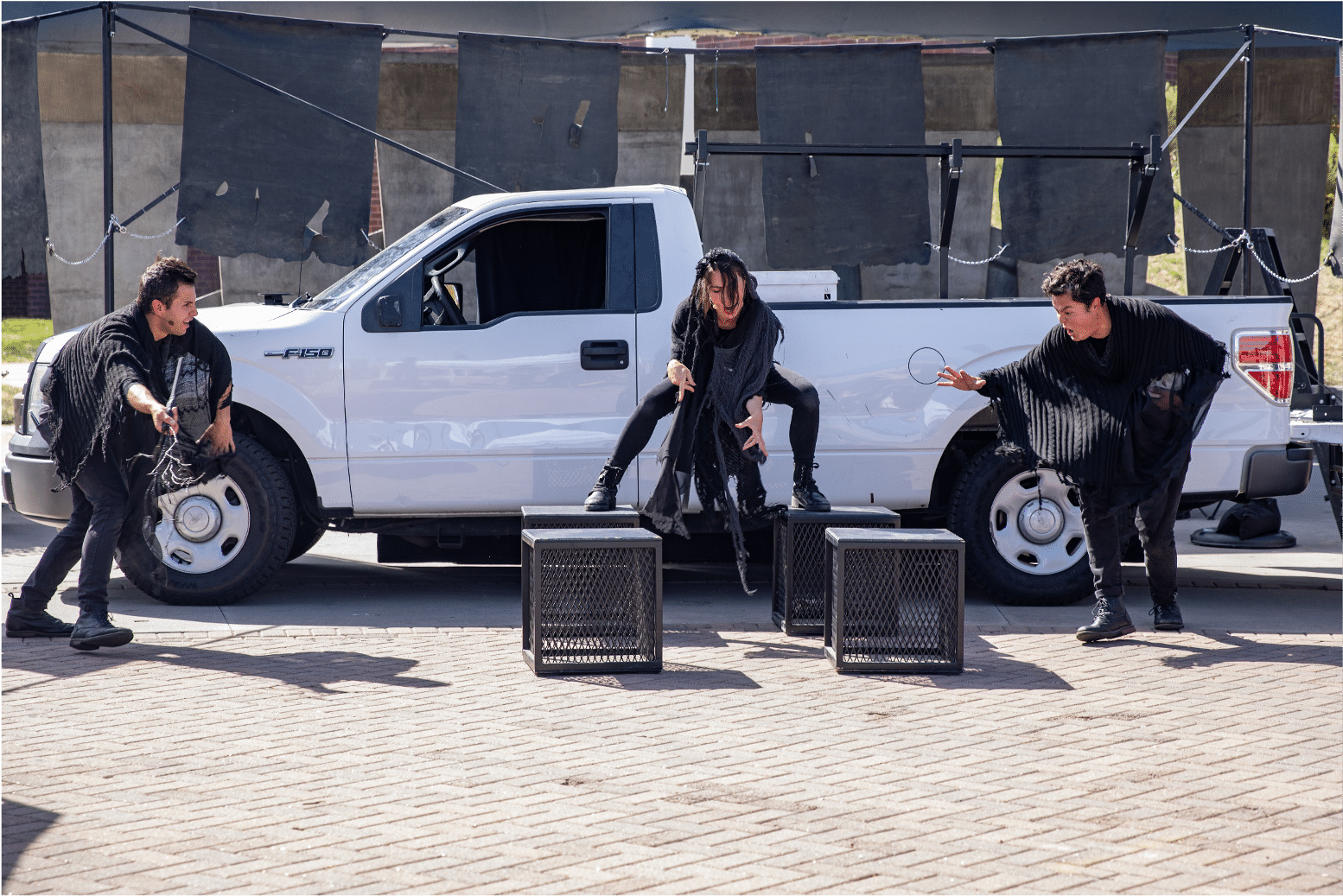 Three actors dressed in black tatters gathered around four-square blocks summoning thin air in front of a white pickup truck